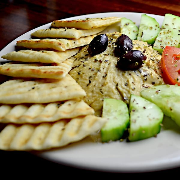 Shawarma Zone's hummus platter with pita bread, olives, cucumber, and tomato on a white plate.